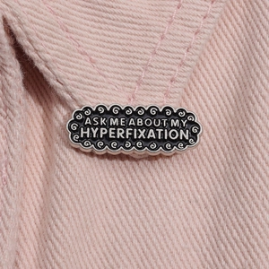 Close-up of black enamel pin with Ask Me About My Hyperfixation text on pink denim