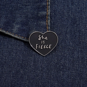 Detail of heart enamel brooch with 'She is FIERCE' text pinned on denim fabric