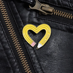 Detail of yellow enamel heart pencil pin with 'MAKE MORE ART' text on black leather jacket