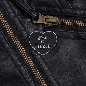 Detailed macro view of a heart-shaped black enamel pin featuring She IS FIERCE on a leather jacket