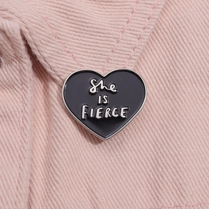 Detail of She IS FIERCE heart enamel pin on pink denim fabric background with metallic edges