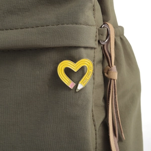 Macro shot of heart-shaped yellow pencil enamel pin with Make More Art text on green bag