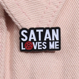 Close-up of Satan Loves Me enamel pin with red pentagram on pink denim fabric background