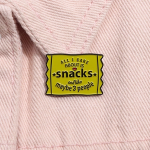Close-up of yellow snack bag enamel pin with black letters and heart on pink fabric