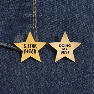 Close-up of gold 5 Star Bitch and silver Doing My Best star pins on blue denim fabric
