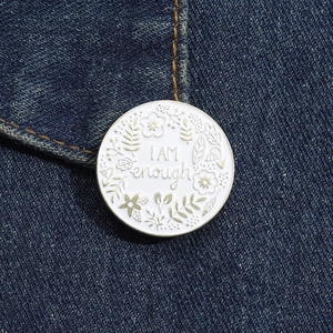 Macro detail of white enamel pin with golden I AM ENOUGH text and floral patterns