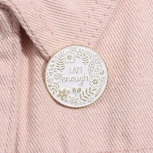 Macro detail of 'I AM enough' white enamel pin with golden floral pattern on pink denim