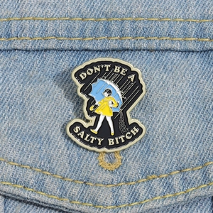 Detail of enamel pin with girl in yellow raincoat and 'Don't be a salty bitch' text