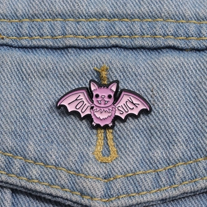 Pink bat enamel brooch with 'YOU SUCK' text pinned on blue denim jacket fabric