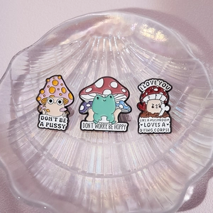 Three unique cartoon mushroom enamel pins displayed on a decorative glass shell tray