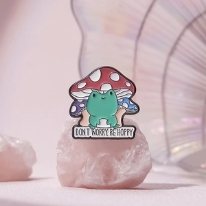 Detail of frog and mushroom enamel pin with "DON'T WORRY, BE HOPPY" text on pink crystals