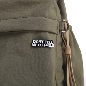 Detail of army green canvas bag featuring a Don't Tell Me To Smile enamel pin badge