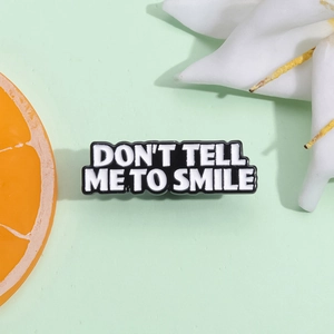 Black enamel pin badge with white text 'Don't Tell Me To Smile' on green background