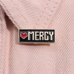 Dettaglio della spilla smaltata Mercy con cuore pixel su denim rosa e bordo dorato
