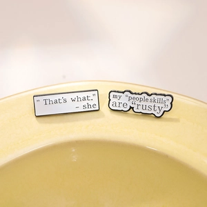 Close-up of set of two enamel pins with funny English quotes on white ceramic dish