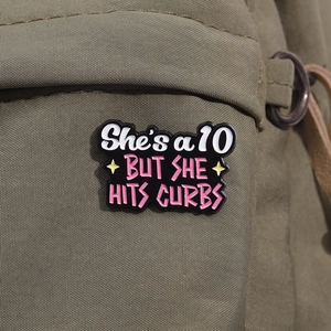 Detail of black enamel pin with She's a 10 text on green canvas backpack texture