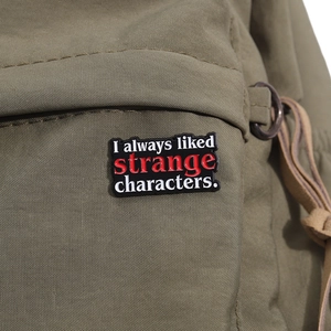 Detail of enamel pin with 'I always liked strange characters' on green canvas fabric