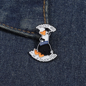 Cartoon graduation duck enamel pin with 'Trust Me I'm a Ducktor' pun on denim background
