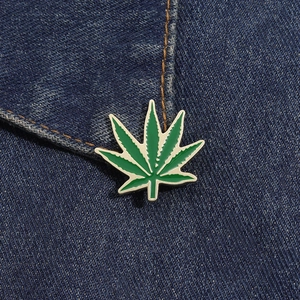 Detailed close-up of a green maple leaf enamel pin pinned onto blue denim fabric