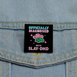 Detail of square frog enamel pin with SLAY-DHD slogan pinned on denim fabric