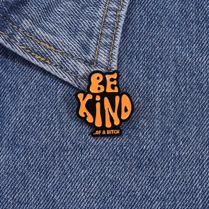 Detailed close-up of a black and orange enamel pin featuring retro Be Kind text on denim