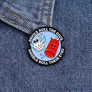 Detail of enamel pin on denim showing a winged dice and cartoon devil motif