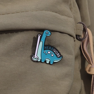 Cute blue dinosaur enamel pin with Herbivore text on a green canvas bag accessory