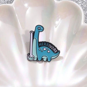 Cartoon blue dinosaur enamel pin with Herbivore text on a decorative white shell tray
