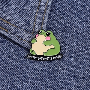Close-up of a cartoon frog enamel pin on denim with Rootin' But Mostly Tootin' text