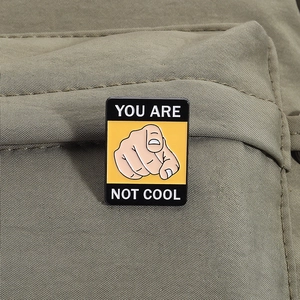 Detail of a rectangular black and yellow enamel pin with YOU ARE NOT COOL text on green fabric