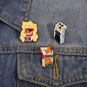 Three cute enamel cat pins with cigarette box themes on a blue denim jacket background