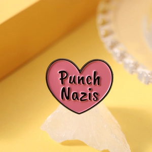 Close-up of pink heart enamel pin with Punch Nazis text on white crystal base