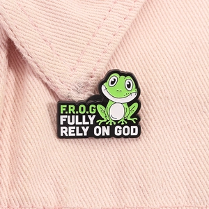 Close-up of green cartoon frog enamel pin with Fully Rely On God text on pink fabric