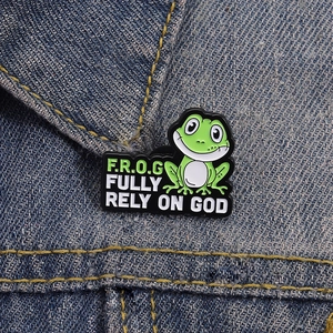Cartoon green frog enamel pin with FULLY RELY ON GOD quote on blue denim jacket