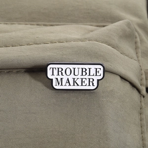 Minimalist white enamel Trouble Maker lapel pin with black text on canvas bag