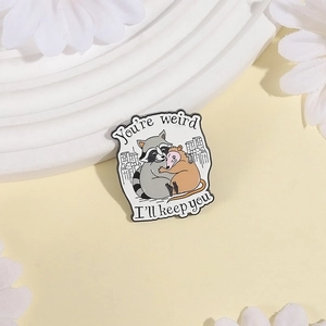 Detail of enamel pin showing a raccoon and opossum hug with text You're weird I'll keep you
