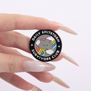 Badge en émail Daily Shitstorm Survivors Club avec nuage et arc-en-ciel en main