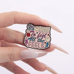Pink enamel pin with It's OKAY To feel All the FEELS text and cartoon icons