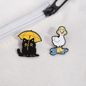 Detail of cute metal enamel pins with black cat holding umbrella and white goose in rain boots