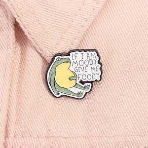 Detail of funny green frog enamel pin with If I Am Moody Give Me Foody text on pink denim