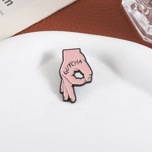 Rosa Emaille-Pin in OK-Handform mit GOTCHA-Schriftzug auf weißem Keramikteller