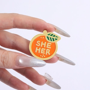 Détail d'un pin's en émail orange avec les pronoms SHE HER tenu dans la main