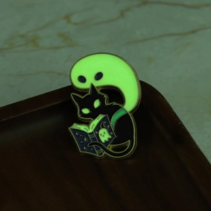 Macro view showing the glowing ghost and black cat reading a magic book on enamel pin