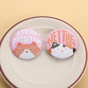 Close-up of cute cat tinplate badges with Ask My Pronouns text on a white ceramic plate
