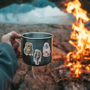 Détail des autocollants de camping à l'aquarelle sur un mug en métal près du feu