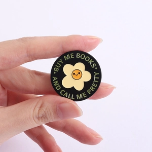 Detail of round enamel pin with flower and 'Buy Me Books' slogan held in hand