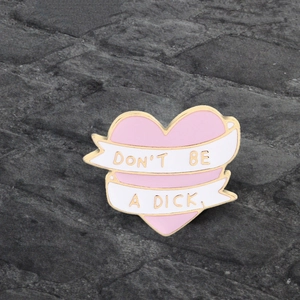 Detail showing pink heart enamel pin with 'DON'T BE A DICK' ribbon and golden metal edges