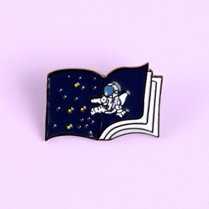 Cartoon astronaut book enamel pin with starry sky design on purple background
