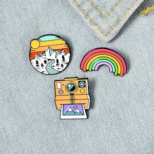Three-piece enamel pin set featuring rainbow, snow mountain, and retro polaroid camera