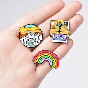 Three enamel pins featuring mountain landscape, retro camera, and colorful rainbow shapes